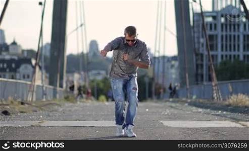 Urban dancer in jeans and sneakers showing some movements over cityscape background.