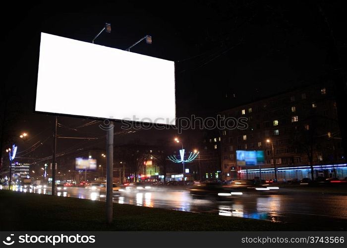Urban scene with an illuminated empty white billboard on the side of a street with cars in motion and a block of flats in the background, by night