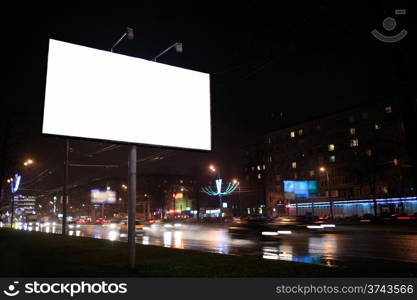 Urban scene with an illuminated empty white billboard on the side of a street with cars in motion and a block of flats in the background, by night