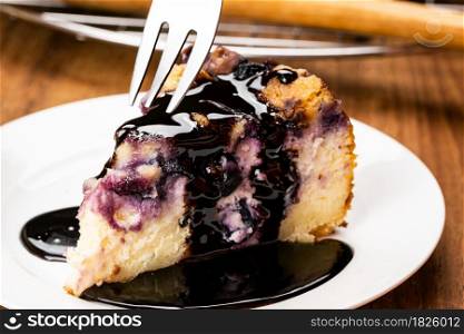 Using metal fork taking a bite from a piece of homemade blueberry and crumble cheesecake topping with chocolate in white ceramic plate on wooden table.