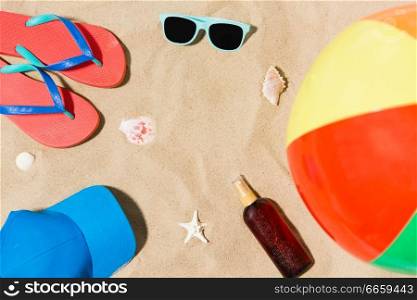 vacation and summer holidays concept - cap, flip flops, sunglasses, beach ball and sunscreen oil with seashells on sand. cap, flip flops and shades and beach ball on sand