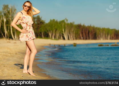 Vacation. Girl in summer dress and sunglasses standing on the empty beach. Young woman relaxing on the sea coast. Summertime.