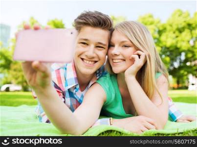 vacation, holidays, technology and friendship concept - smiling couple lying on blanket and making selfie with smartphone in park
