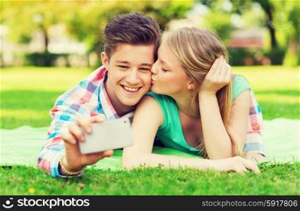 vacation, holidays, technology, love and friendship concept - kissing couple lying on blanket and making selfie with smartphone in park