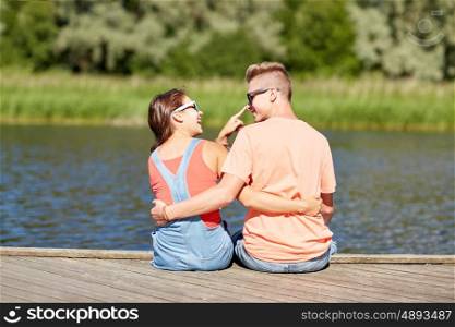 vacation, love and people concept - happy teenage couple sitting and hugging on river berth at summer