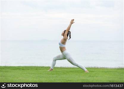 Vacation of Attractive Asian woman relaxing in yoga Warrior one pose on green grass and beach with sunset beautiful sea in Tropical island,Feeling comfortable and relax in holiday,Vacations Concept