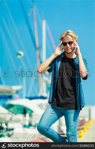 Vacation, relaxation concept. Young fashionable blonde man standing near harbor, relaxing and enjoying beautiful, sunny weather. Outdoor shot. Blonde man standing near harbor in summer