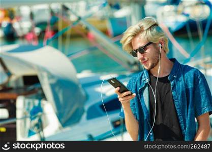 Vacation, relaxation, passion concept. Young fashionable blonde in sunglasses man relaxing, listening to music and enjoying beautiful, sunny weather. Outdoor shot. Blonde man in sunglasses listening to music
