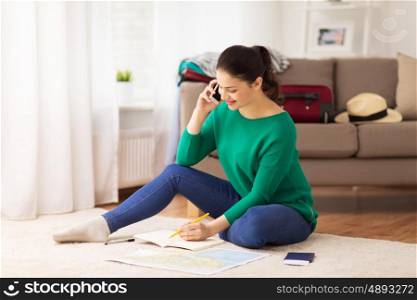 vacation, tourism, travel and people concept - happy young woman with notebook, passport and map calling on smartphone at home