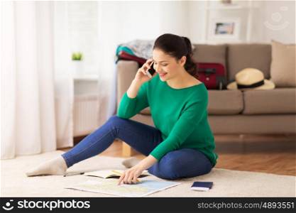vacation, tourism, travel and people concept - happy young woman with notebook, passport and map calling on smartphone at home
