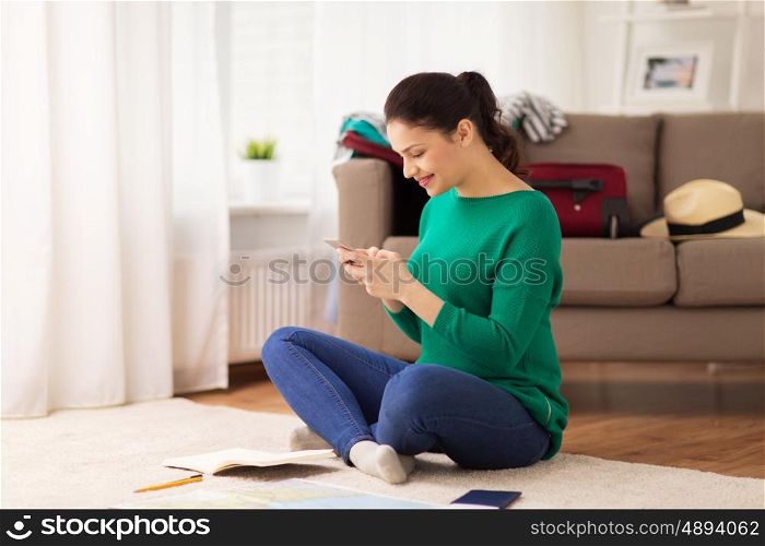 vacation, tourism, travel and people concept - happy young woman with notebook, passport and map texting on smartphone at home