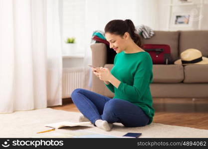 vacation, tourism, travel and people concept - happy young woman with notebook, passport and map texting on smartphone at home