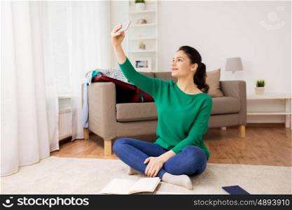 vacation, tourism, travel and people concept - happy young woman with smartphone going on trip and taking selfie at home