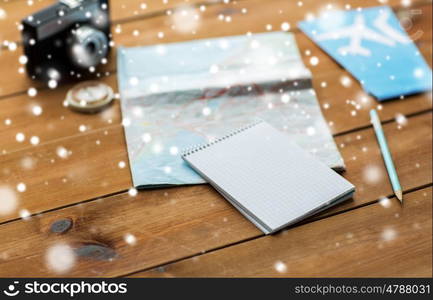 vacation, tourism, travel and winter holidays concept - notepad with map, camera and airplane tickets over snow