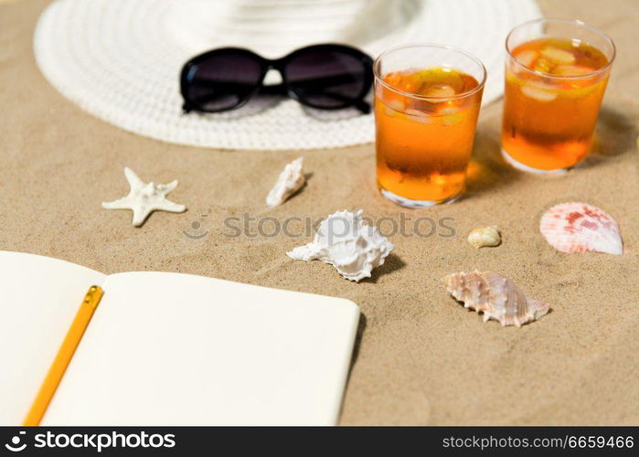 vacation, travel and summer concept - notebook with pencil, two glasses of aperitif cocktails, sun hat and sunglasses on beach sand. notebook, cocktails, hat and shades on beach sand