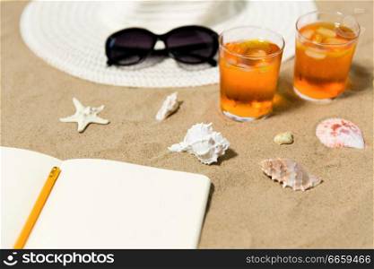 vacation, travel and summer concept - notebook with pencil, two glasses of aperitif cocktails, sun hat and sunglasses on beach sand. notebook, cocktails, hat and shades on beach sand