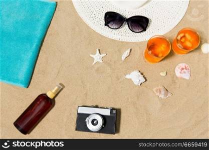 vacation, travel and summer concept - two glasses of aperitif cocktails with ice, sun hat with sunglasses, vintage film camera with sunscreen oil and seashells on beach sand. drinks, hat, camera and sunglasses on beach sand