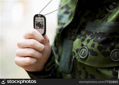 vaccination, army and pandemic concept - close up of soldier in military uniform holding dog tag with qr code. close up of young soldier with qr code on dog tag