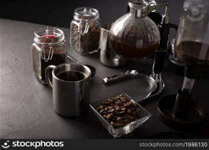 Vacuum coffee maker also known as vac pot, siphon or syphon coffee maker. Metallic cup and toasted coffee beans on rustic black stone table.