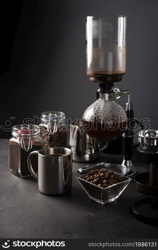 Vacuum coffee maker also known as vac pot, siphon or syphon coffee maker. Metallic cup and toasted coffee beans on rustic black stone table