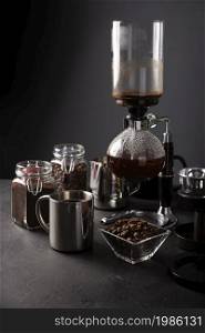 Vacuum coffee maker also known as vac pot, siphon or syphon coffee maker. Metallic cup and toasted coffee beans on rustic black stone table