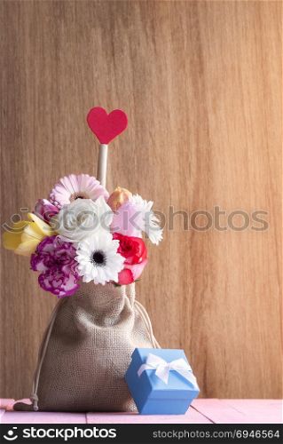 Valentine day card with a beautiful bouquet of flowers in a jute bag, with a red heart and a blue gift box, on a pink table and a wooden background.