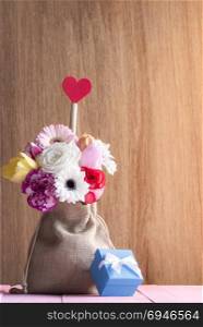 Valentine day card with a beautiful bouquet of flowers in a jute bag, with a red heart and a blue gift box, on a pink table and a wooden background.
