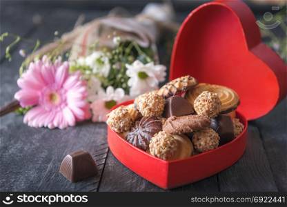 Valentine day theme image with a heart-shaped gift box full of cookies and chocolates, a lovely bouquet of flowers in the background, on a black table.