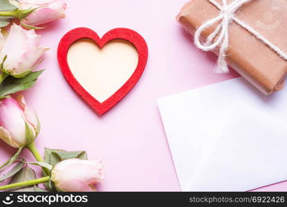 Valentine day theme image with a red heart with copy space in the middle, surrounded by pink roses, an envelope and a gift wrapped in brown paper, on a pink background.