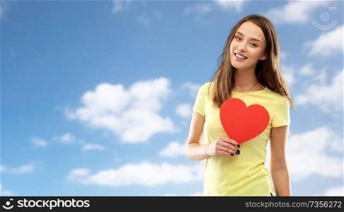 valentine&rsquo;s day and people concept - smiling young woman or teenage girl in yellow t-shirt with red heart over blue sky and clouds background. smiling teenage girl with red heart over sky