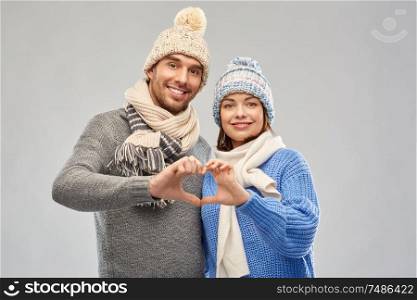 valentine&rsquo;s day, christmas and love concept - happy romantic couple in knitted hats and scarves making hand heart gesture over grey background. happy couple in winter clothes making hand heart