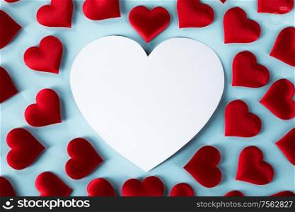 Valentine&rsquo;s day many red silk hearts and white heart shaped card on blue background, love concept. Valentines day hearts on blue