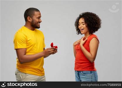 valentine&rsquo;s day, proposal and couple concept - smiling african american man giving diamond engagement ring in little red box to happy woman over grey background. african american man giving woman engagement ring