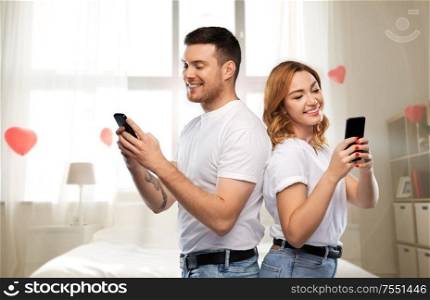 valentine&rsquo;s day, technology and people concept - happy couple in white t-shirts with smartphones over bedroom decorated with heart shaped balloons background. happy couple with smartphones on valentines day
