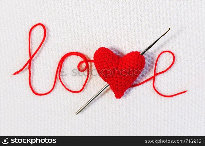 Valentine&rsquo;s day, wedding backdrop. Crochet hook pierced red knitted volume heart like an arrow in word love from threads on white knitted background. Template for design, valentines card, invitation.. Valentine&rsquo;s day, wedding backdrop. Crochet hook pierced red knitted volume heart like an arrow in word love from threads on white knitted background. Template for design, valentines card, invitation