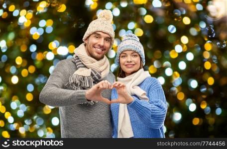 valentine&rsquo;s day, winter holidays and love concept - happy romantic couple in knitted hats and scarves making hand heart gesture over christmas lights background. happy couple in winter clothes making hand heart
