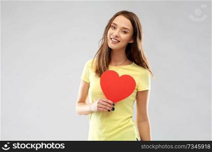 valentine’s day and people concept - smiling young woman or teenage girl in yellow t-shirt with red heart over grey background. smiling teenage girl with red heart