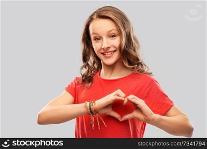 valentine’s day, love and charity concept - smiling teenage girl with long hair in red t-shirt making hand heart gesture over grey background. smiling teenage girl in red making hand heart