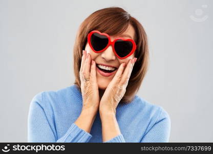 valentine’s day, summer and old people concept - portrait of smiling senior woman in red heart-shaped sunglasses over grey background. smiling senior woman in heart-shaped sunglasses