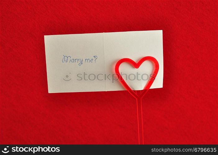 Valentines day. A note holder with a red heart with a note reading marry me isolated against a red background