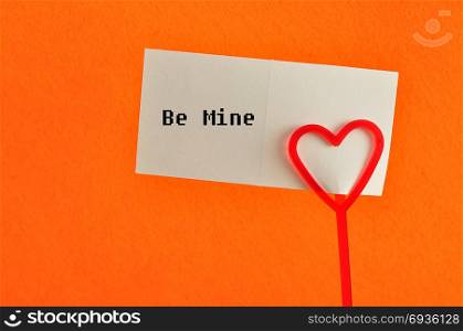 Valentines day. A note holder with a red heart with a note reading be mine isolated against an orange background