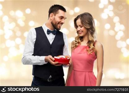 valentines day and people concept - happy couple with chocolate box in shape of heart over beige background and festive lights. happy couple with chocolate box in shape of heart