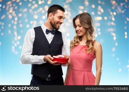 valentines day and people concept - happy couple with chocolate box in shape of heart over holiday lights on blue background. happy couple with chocolate box in shape of heart