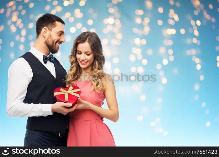 valentines day and people concept - happy couple with gist box in shape of heart over holiday lights on blue background. happy couple with gift on valentines day