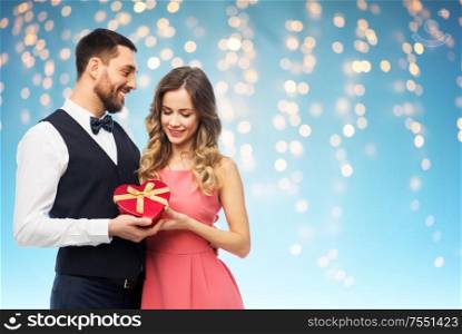 valentines day and people concept - happy couple with gist box in shape of heart over holiday lights on blue background. happy couple with gift on valentines day