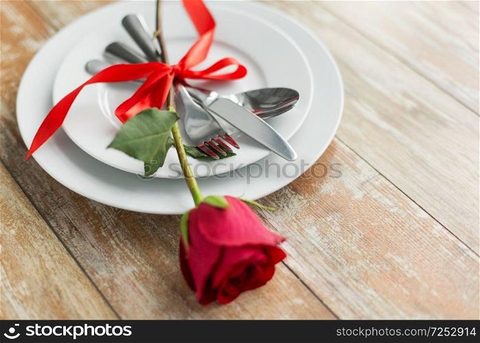 valentines day and romantic dinner concept - close up of red rose flower on set of dishes with cutlery on wooden table. close up of red rose flower on set of dishes