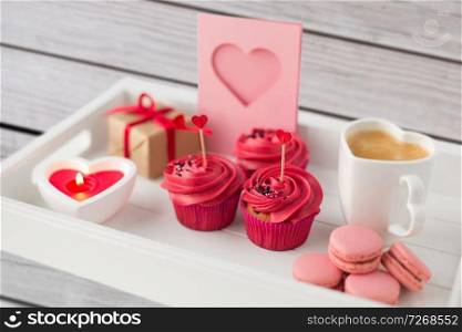 valentines day and sweets concept - close up of cupcakes with red buttercream frosting and heart shaped cocktail sticks, macarons, candle, coffee cup and gift box on tray. close up of red sweets for valentines day
