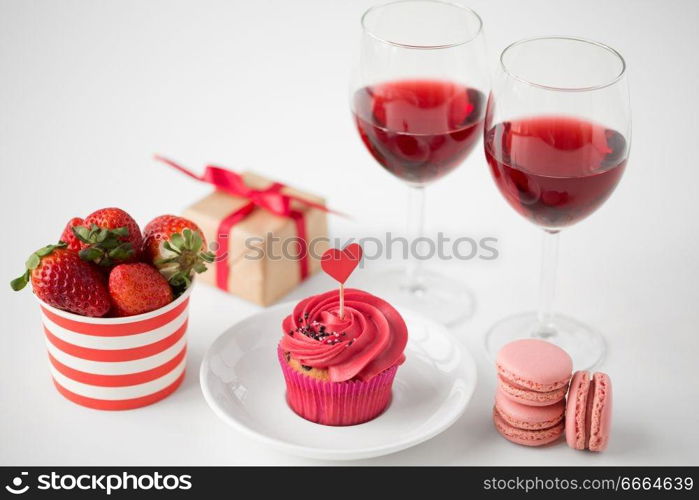 valentines day and sweets concept - close up of frosted cupcake with red heart shaped cocktail stick, macarons, strawberries, glasses of wine and gift box on white background. close up of red sweets for valentines day