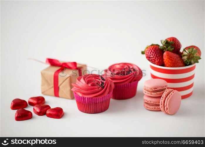 valentines day and sweets concept - close up of frosted cupcakes, red heart shaped chocolate candies, macarons, strawberries and gift box on white background. close up of red sweets for valentines day