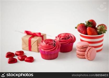 valentines day and sweets concept - close up of frosted cupcakes, red heart shaped chocolate candies, macarons, strawberries and gift box on white background. close up of red sweets for valentines day
